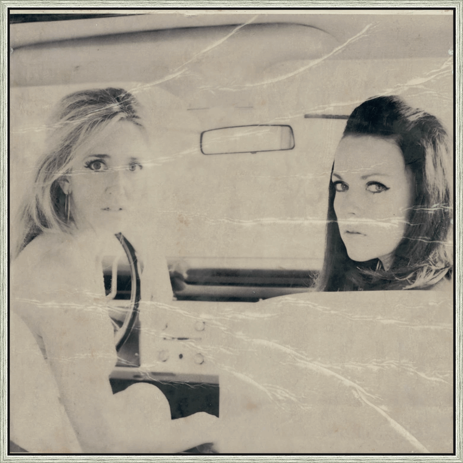 This vintage black and white print captures a moment of timeless elegance inside a car. Two women with striking features, and dramatic eye makeup, are framed within the vehicle's interior, their gazes directed towards the viewer. The slightly distressed paper adds a layer of nostalgic charm, suggesting a snapshot from a bygone era.