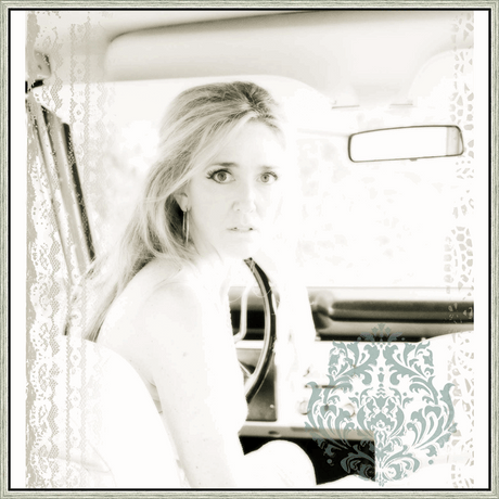 A captivating black and white image captures a woman sitting inside of a car, gazing directly at the viewer with a look of intrigue. Soft focus and a delicate aesthetic gives the piece a vintage quality. The artwork's monochrome treatment enhances the emotional depth of the scene, while decorative embellishments provide a sense of timeless elegance.