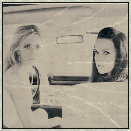 This vintage black and white print captures a moment of timeless elegance inside a car. Two women with striking features, and dramatic eye makeup, are framed within the vehicle's interior, their gazes directed towards the viewer. The slightly distressed paper adds a layer of nostalgic charm, suggesting a snapshot from a bygone era.